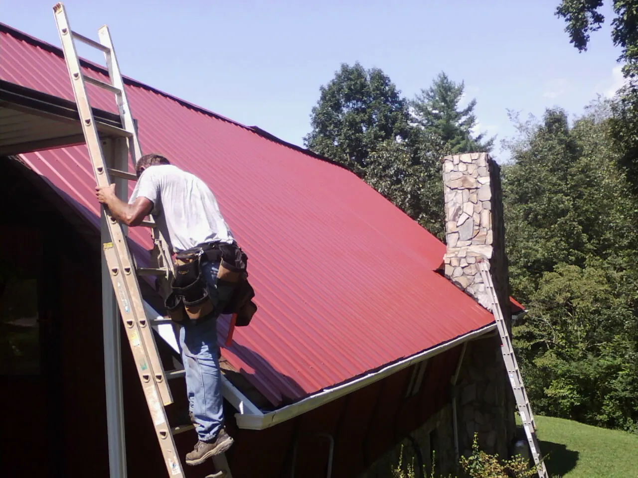Roofer on a ladder beside a red metal roof and stone chimney — Soffit & Fascia Repair by Roofing Brothers in Lakeland