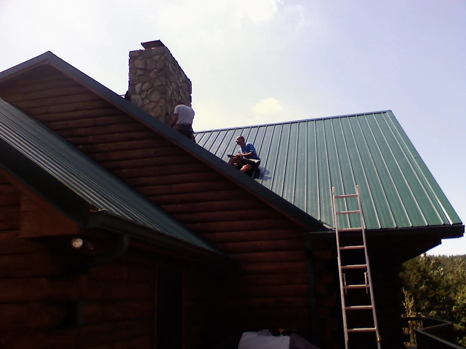 Roofers installing green metal panels beside a stone chimney on a log home in Lakeland